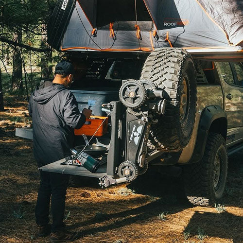 Man using a mounted 2-ton "The Beast" Jack 2.0 on an SUV in a forest campground.